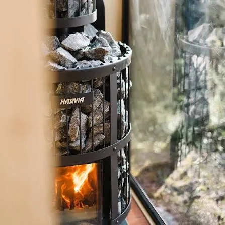 Harvia wood-fired heater stacked with stones, fire glowing in the firebox beside a sauna window